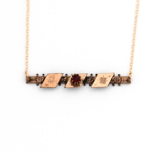 Puede incluir: Un collar de oro con un colgante que presenta una fila de tres dijes en forma de diamante. El dije central tiene una piedra preciosa roja, mientras que los otros dos dijes tienen diseños florales.