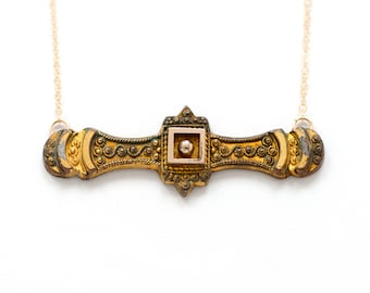 Puede incluir: Un collar dorado de estilo vintage con un colgante de barra ornamentado. El colgante presenta detalles intrincados, un engaste cuadrado central con un pequeño acento redondo, y está suspendido de una delicada cadena dorada. El collar tiene una estética clásica y antigua.