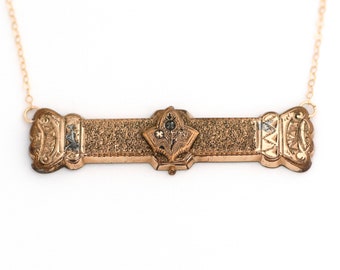 Puede incluir: Un collar de oro con un colgante rectangular largo que presenta un diseño floral con un pequeño diamante en el centro. El colgante tiene una superficie texturizada y bordes ornamentados.