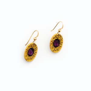 May include: A pair of gold earrings with a purple gemstone in the centre of each earring. The earrings have a detailed, ornate design.