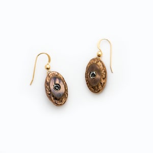 May include: A pair of gold earrings with oval-shaped bronze pendants. Each pendant has a textured design and a small, clear gemstone in the centre.