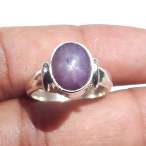 May include: A silver ring featuring a large, oval-shaped, purplish-pink gemstone. The ring has a simple band with decorative accents on either side of the gemstone setting. The gemstone has a star-like appearance.