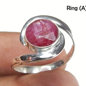 Puede incluir: Un anillo de plata con una gran gema de rubí redonda y facetada. La banda del anillo se curva alrededor del dedo, con la gema engastada en un bisel. El texto "Ring (A)" es visible.