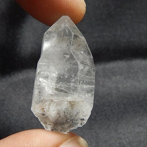 Peut inclure: Une pointe de cristal de quartz naturel transparent avec une terminaison pointue et une structure prismatique hexagonale. Le cristal présente des fractures internes, des stries sur ses faces verticales et une base trouble et texturée.