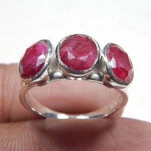 Puede incluir: Un anillo de plata con tres rubíes ovalados y facetados. Los rubíes son de un color rojo intenso y están engastados en biseles individuales. El anillo tiene una banda lisa y pulida.