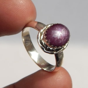 May include: A silver ring featuring a large, oval-shaped, purplish-red gemstone. The gemstone is set in a decorative, scalloped bezel. The ring is held by a finger against a plain background.