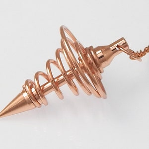 Puede incluir: Un péndulo de metal de color oro rosa con un diseño en espiral y una punta puntiaguda. El péndulo está unido a una cadena.