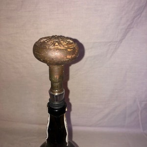 Może przedstawiać: Antyczny korek do butelki z ozdobnym metalowym wierzchołkiem i ciemnobrązową szklaną butelką.