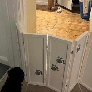 May include: A white folding pet gate featuring four rectangular panels connected by metal hinges. Each panel is decorated with dark grey paw print decals. The gate is positioned to block a doorway, standing upright on a floor surface.