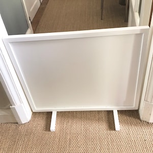 May include: White freestanding baby gate with a white frame and two white legs. The gate is in front of a doorway with a brown carpet.