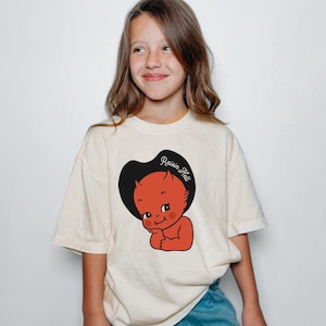 May include: Cream-colored t-shirt featuring a cartoon devil wearing a black cowboy hat with the text "Raisin Hell". The devil is red with a cute expression. The shirt is paired with blue jeans.