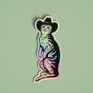 May include: A sticker of a cat wearing a black cowboy hat. The cat is illustrated in black and white with striped markings, and the sticker has a holographic, iridescent border. The cat is sitting upright.