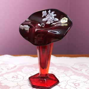 May include: A ruby red glass vase with a flared top and a hexagonal base. The vase features a white floral design on the top and a sticker. The vase is approximately 10 inches tall.
