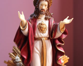 Może przedstawiać: Religijna figurka Jezusa Chrystusa, z wyciągniętymi ramionami. Posąg ma białą szatę, czerwony płaszcz ze złotymi wykończeniami i złotą aureolę. Złote serce jest widoczne na piersi. Posąg ma około 30 cm wysokości.