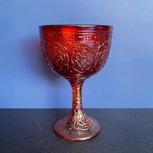 May include: A ruby red, ornate glass goblet with a detailed floral pattern. The stem and base are also decorated with embossed designs. The glass has a glossy finish and is set against a blue background.