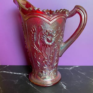 May include: A red iridescent glass pitcher with a floral design. The pitcher has a scalloped rim and a handle.