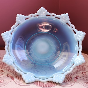 May include: A light blue, opalescent glass bowl with a scalloped edge and shell-shaped accents. The bowl has a round, open center and sits on a white lace tablecloth. The bowl's rim has a delicate, openwork design.
