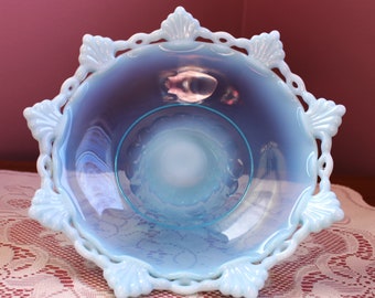 May include: A light blue, opalescent glass bowl with a scalloped edge and shell-shaped accents. The bowl has a round, open center and sits on a white lace tablecloth. The bowl's rim has a delicate, openwork design.