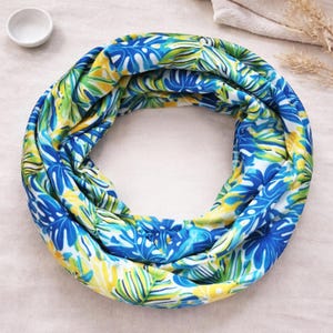 May include: A circular infinity scarf featuring a vibrant tropical leaf pattern in shades of bright blue, lime green, and yellow. The fabric is arranged in a continuous loop, showcasing a dense, repeating botanical print across the entire surface.