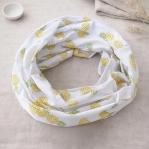 May include: A white infinity scarf featuring a repeating pattern of yellow pineapples with green leaves. The fabric is draped in a circular shape, showcasing the consistent pineapple print across the soft, lightweight material of the accessory.