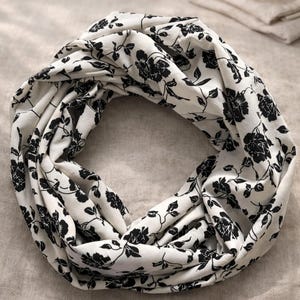 May include: An infinity scarf featuring a white base colour with a repeating black floral pattern of roses and leaves. The fabric is arranged in a circular, twisted loop, showcasing the continuous print across the entire surface of the accessory.