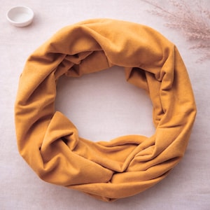 May include: A mustard yellow infinity scarf made of a soft, matte fabric. The scarf is arranged in a circular, draped loop, showcasing its continuous, seamless design and rich, solid colour throughout the entire piece of fabric.