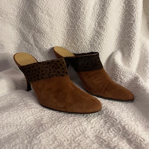 May include: A pair of brown suede heeled mules with a leopard print detail on the back of the heel.