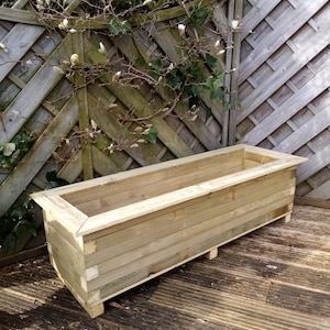 May include: A long, rectangular wooden planter box with a natural finish. The planter is made of multiple wooden planks and has a raised lip around the top. The planter is sitting on a wooden deck.