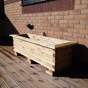 May include: A wooden planter box with a natural finish sits on a wooden deck. The planter box is made of multiple pieces of wood that are stacked on top of each other. The planter box is sitting on a wooden deck that is made of multiple pieces of wood that are stacked on top of each other.