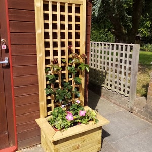 May include: A wooden planter with a trellis attached to the back. The planter is filled with green plants and pink flowers. The planter is sitting in front of a wooden shed.