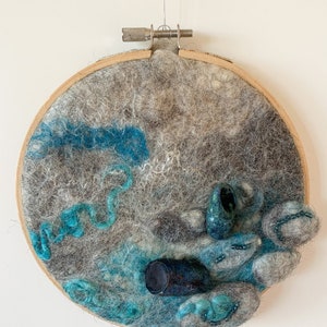 May include: A felted art piece in a wooden embroidery hoop. The artwork features a textured landscape with gray and teal wool, depicting abstract shapes and forms. Small, dark blue cylindrical and rounded objects are incorporated.