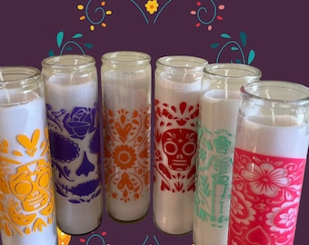May include: A collection of seven glass jar candles with white wax. Each candle features a different colorful Day of the Dead-themed design, including skulls, flowers, and decorative patterns in orange, purple, red, yellow, and pink.