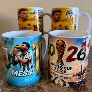 Könnte beinhalten: Vier weiße Keramiktassen mit Fußballmotiven. Zwei Tassen zeigen Porträts der Spieler James Rodriguez und Luis Diaz in gelben Trikots. Eine Tasse zeigt Lionel Messi, wie er den Weltpokal küsst. Eine Tasse zeigt das Logo der FIFA Fußball-Weltmeisterschaft 2026.