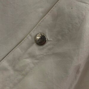 May include: Close-up of white fabric with a silver button. The button is embossed with the text "COURTURE" and a logo. The fabric is creased and has visible stitching.