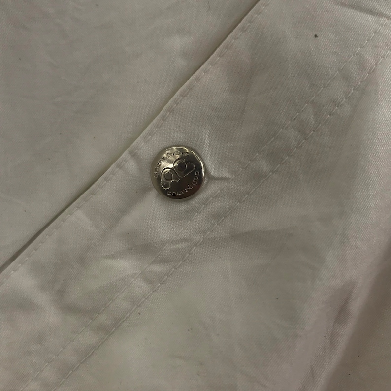 May include: Close-up of white fabric with a silver button. The button is embossed with the text "COURTURE" and a logo. The fabric is creased and has visible stitching.