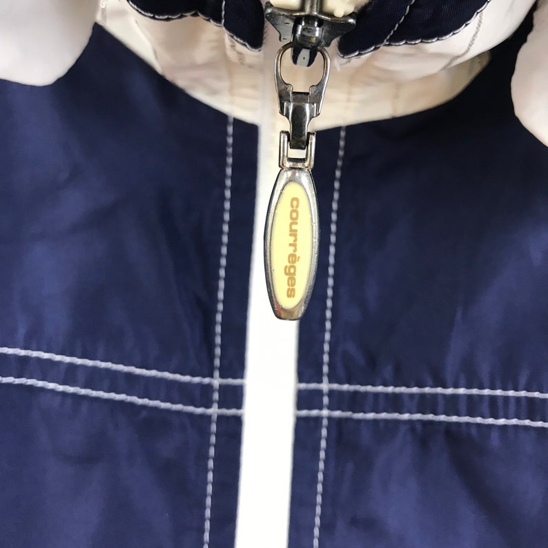 May include: Close-up of a jacket zipper with the brand name "courr&egrave;ges" on the pull tab. The jacket is navy blue and white with white stitching. The zipper is silver.