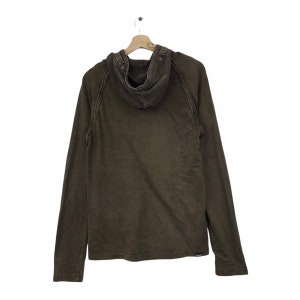 May include: A brown hooded long-sleeve shirt hanging on a wooden hanger. The shirt has a buttoned hood and a small logo near the bottom hem. The fabric appears to be a textured cotton or similar material.