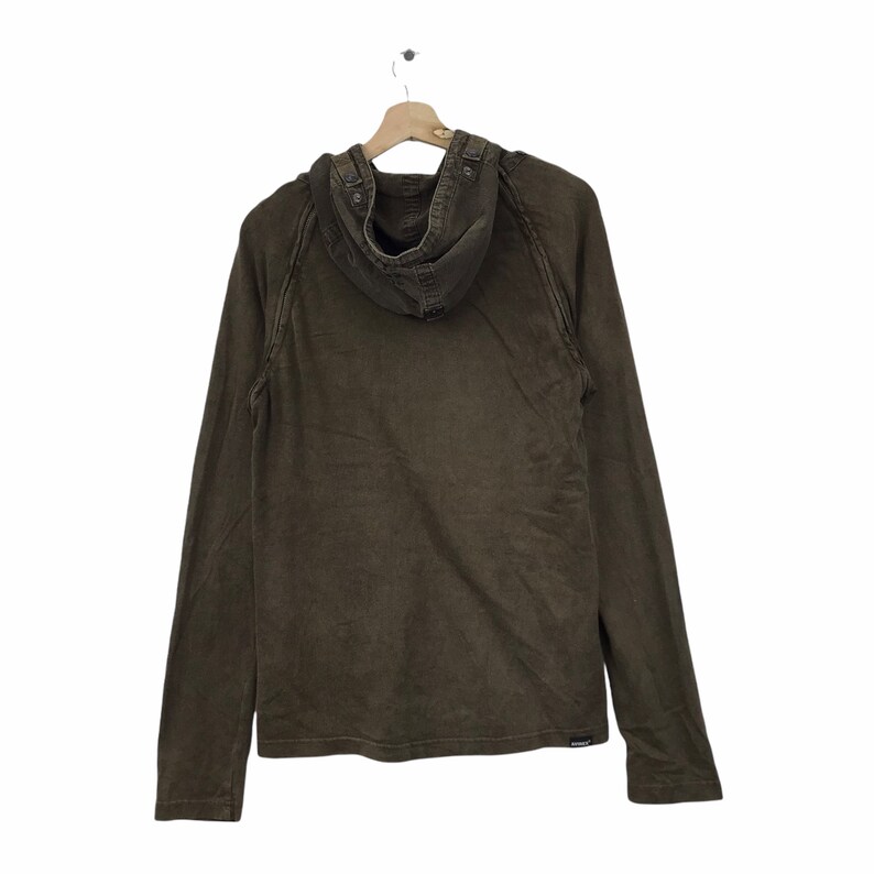 May include: A brown hooded long-sleeve shirt hanging on a wooden hanger. The shirt has a buttoned hood and a small logo near the bottom hem. The fabric appears to be a textured cotton or similar material.