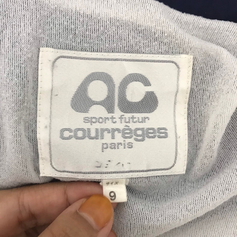 May include: Close-up of a white label with the text "AC sport futur courr&egrave;ges paris". The label also has the number "9" and other Japanese text. The label is on a white and navy blue fabric.