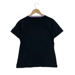 May include: Black t-shirt with a purple collar, hanging on a wooden hanger. The short-sleeved shirt is made of a soft material. The back of the shirt is visible, showing the collar and the shape of the garment.
