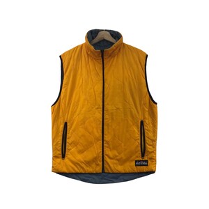 May include: A mustard yellow quilted vest featuring a high collar, a full-length black front zipper, and two black zippered side pockets. The garment includes a black rectangular patch with the white text Wild Things on the lower right side.