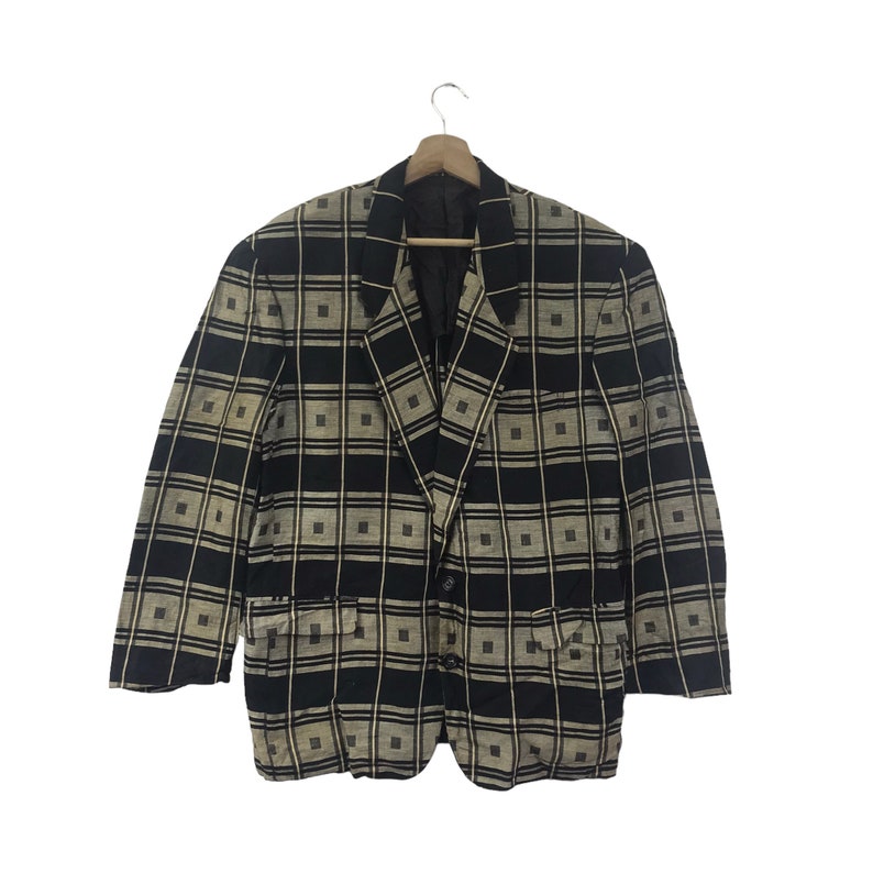 May include: A black and tan plaid blazer with a square pattern. The blazer has two front pockets and a two-button closure. The jacket is hanging on a wooden hanger.