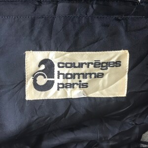 May include: Close-up of a label with the text "courr&egrave;ges homme paris" in black font on a cream-colored background. The label is sewn onto a dark blue, textured fabric. The Courr&egrave;ges logo is a stylized "a".