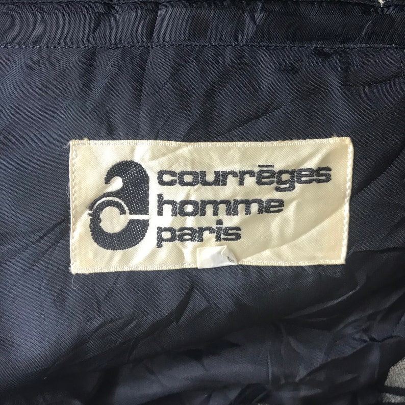 May include: Close-up of a label with the text "courr&egrave;ges homme paris" in black font on a cream-colored background. The label is sewn onto a dark blue, textured fabric. The Courr&egrave;ges logo is a stylized "a".