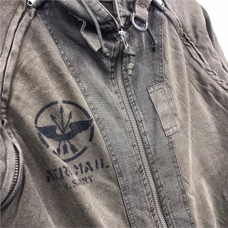 May include: Close-up of a military-style olive green jacket with a full zipper and a hood. The jacket features a black graphic with the words "AIR MAIL U.S. ARMY" printed below the graphic. The jacket has a distressed, worn look.