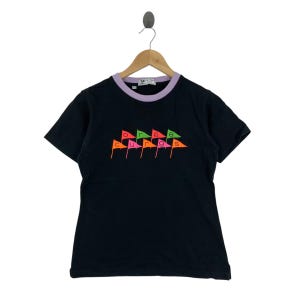 May include: Black t-shirt with a purple and white collar. The shirt features the word "courage" in colorful flag-shaped letters. The shirt is hanging on a wooden hanger.