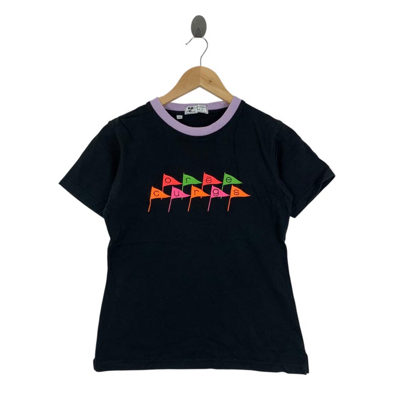 May include: Black t-shirt with a purple and white collar. The shirt features the word "courage" in colorful flag-shaped letters. The shirt is hanging on a wooden hanger.