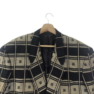 May include: A black and beige plaid blazer hanging on a wooden hanger. The blazer has a classic design with a notched lapel and a two-button closure. The plaid pattern is composed of black and beige squares and rectangles.