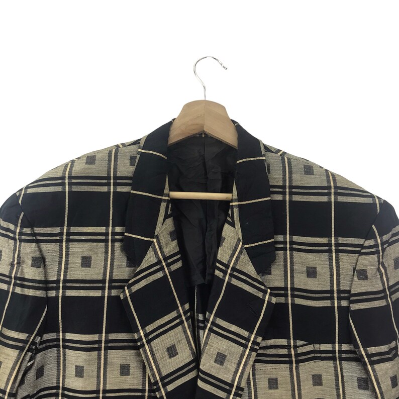 May include: A black and beige plaid blazer hanging on a wooden hanger. The blazer has a classic design with a notched lapel and a two-button closure. The plaid pattern is composed of black and beige squares and rectangles.