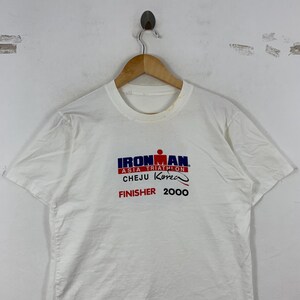 K&ouml;nnte beinhalten: Ein wei&szlig;es T-Shirt mit kurzen &Auml;rmeln und einem Aufdruck auf der Brust mit der Aufschrift IRONMAN ASIA TRIATHLON CHEJU Korea FINISHER 2000. Der Text ist in Blau, Rot und Schwarz gedruckt, wobei das Ironman-Logo &uuml;ber den Veranstaltungsdetails steht.